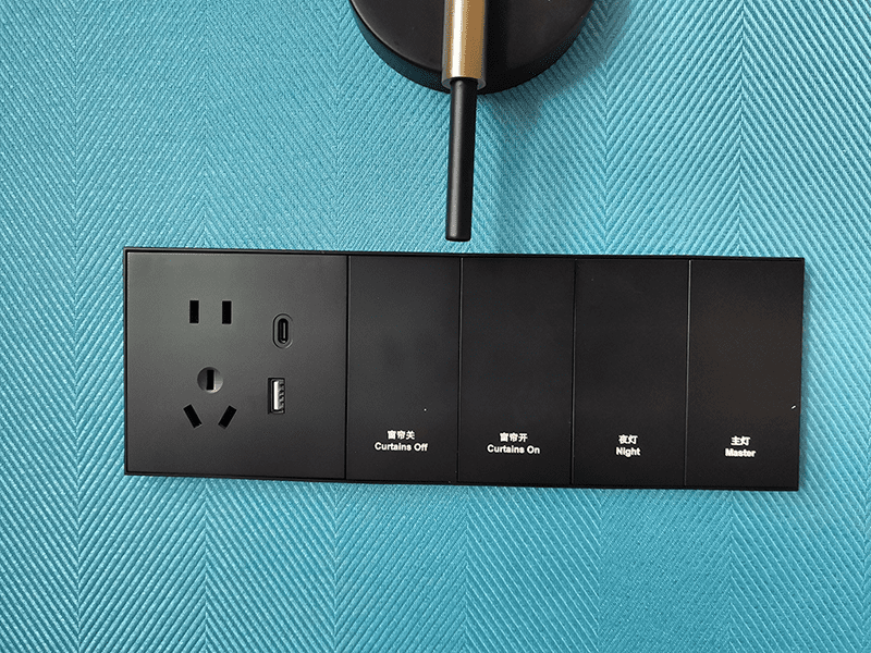 On-site installation photo of black eco painted plastic Zigbee smart switch panel for Hilton hotel project, integrated with 5-hole power socket, USB & Type-C charging port, 4 control buttons for master light, night light, curtain open and curtain close, mounted on blue textured wall fabric